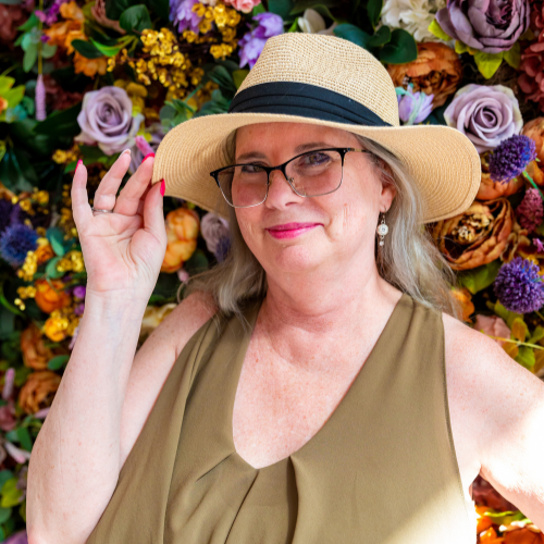 female with glasses and hat in front of wall of flowers