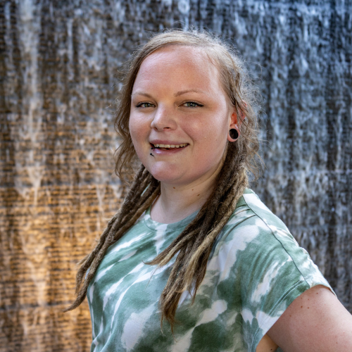 female with dreads smiling standing in front of a wall of water flowing down