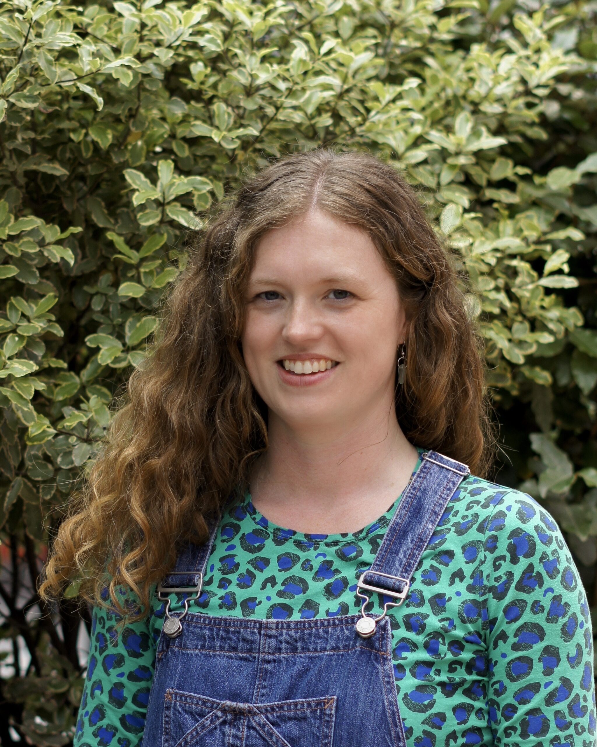 female smiling standing in front of a tree with overalls on